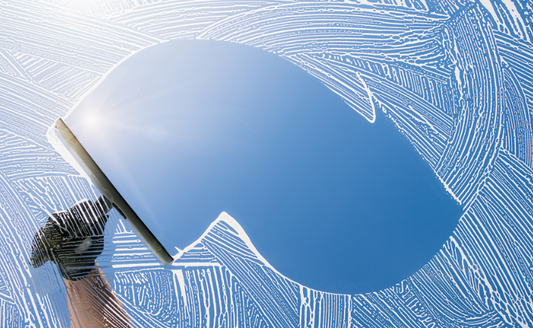 Glass and window cleaner using squeegee to create clean arc through soapy window, revealing bright blue sky